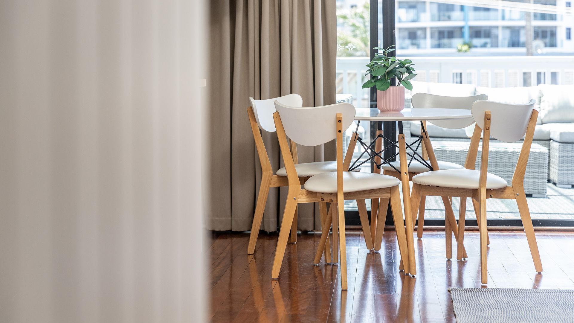 Un coin repas avec quatre chaises blanches et une table ronde, une plante en pot, et une vue sur un bâtiment à travers de grandes fenêtres.