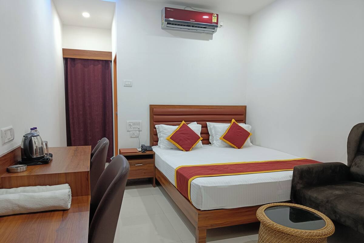 A hotel room with a wooden bed, a desk with a kettle, a chair, a bedside table, and a wicker basket.