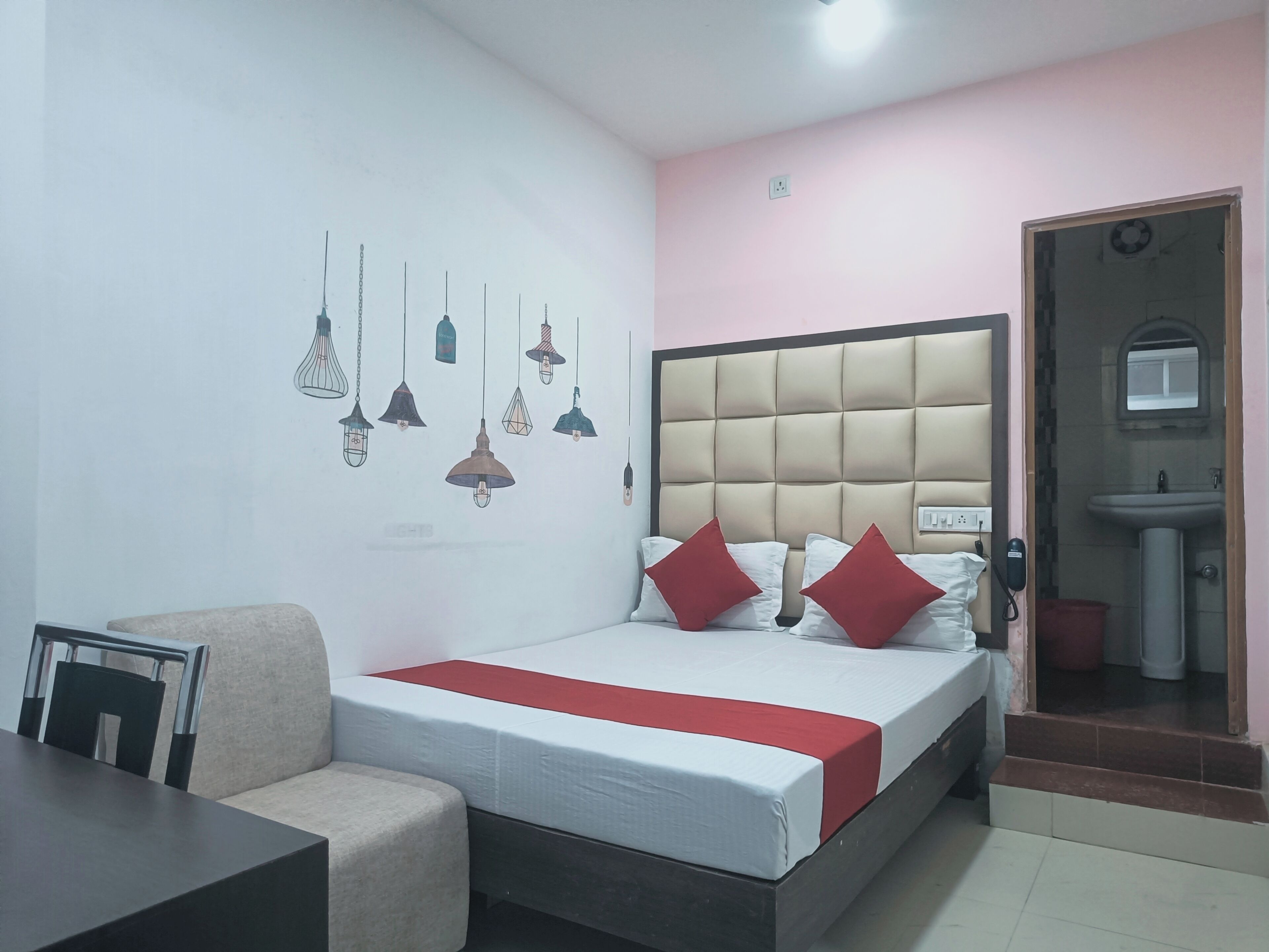 A bedroom with a bed, bedside table, chair, hanging light fixtures, and a bathroom with a sink and mirror.