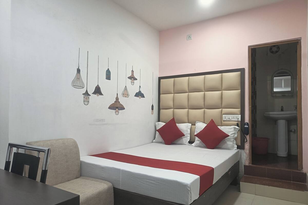A hotel room with a bed, a bedside table, a chair, a wall-mounted light fixture, and a bathroom visible through an open door.