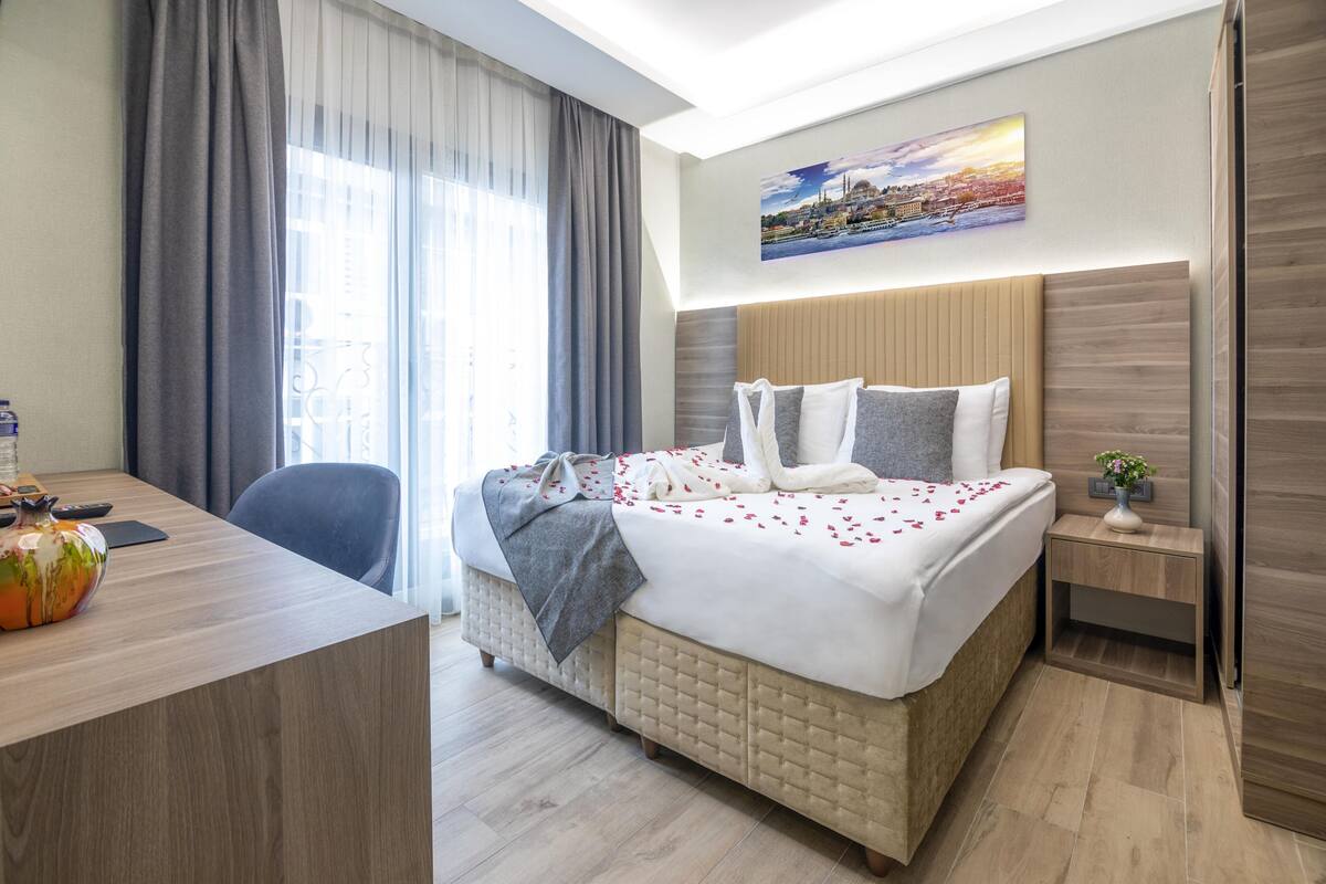A hotel room with a bed, a desk, a chair, a nightstand, and a painting on the wall.