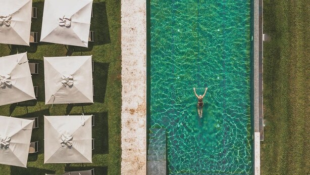 Vista aérea de una piscina rectangular con agua cristalina, rodeada por una solana de piedra y varias sombrillas blancas.
