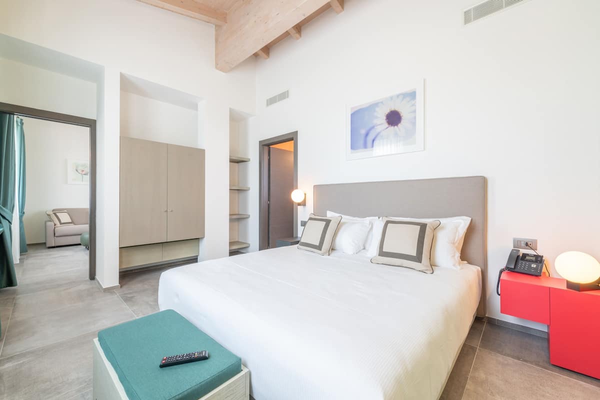 Ein Hotelzimmer mit einem Bett, einem roten Nachttisch, einer Fernbedienung auf einem grünen Hocker und einem Gemälde an der Wand.