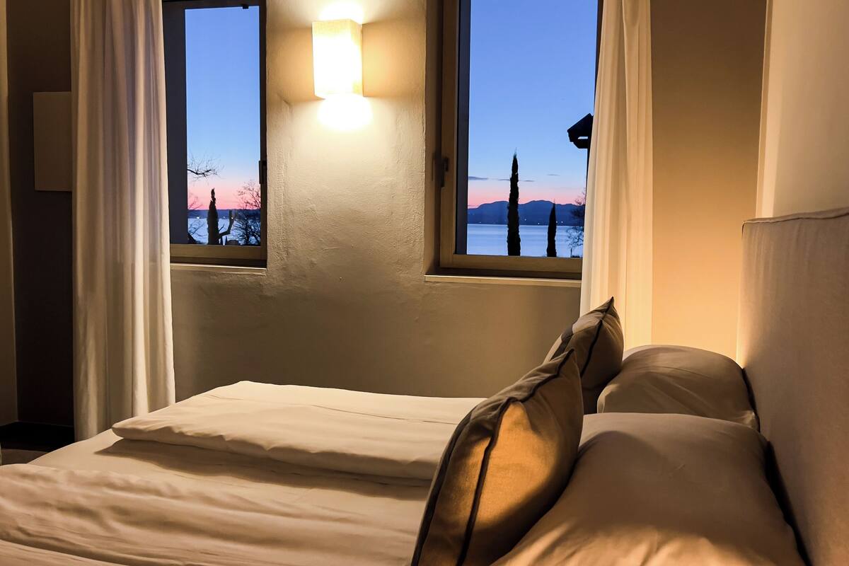 Ein Bett mit weißer Bettwäsche und Kissen, ein Nachttisch und ein Fenster mit Blick auf Bäume und einen Sonnenuntergang.