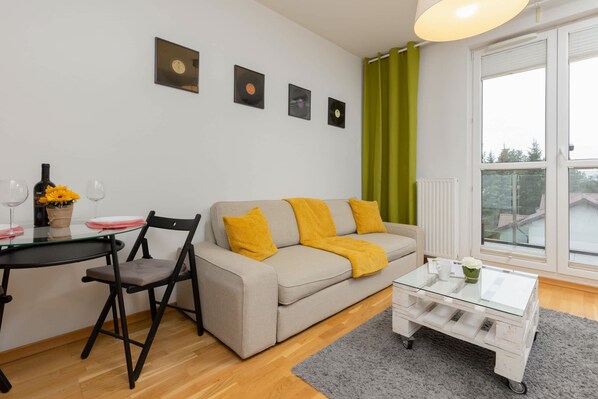 A modern living room with a sofa, a glass coffee table, a dining table with chairs, and a window with curtains. - Bright Apartment with Balcony | 15 min to Metro (Warszawa)