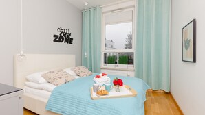 Un dormitorio con cama, ventana con cortinas, pared con un letrero y una mesita de noche con objetos sobre la cama.