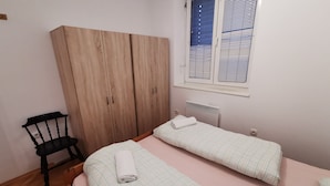 A bedroom with two single beds, a wooden wardrobe, a chair, and a window with blinds.