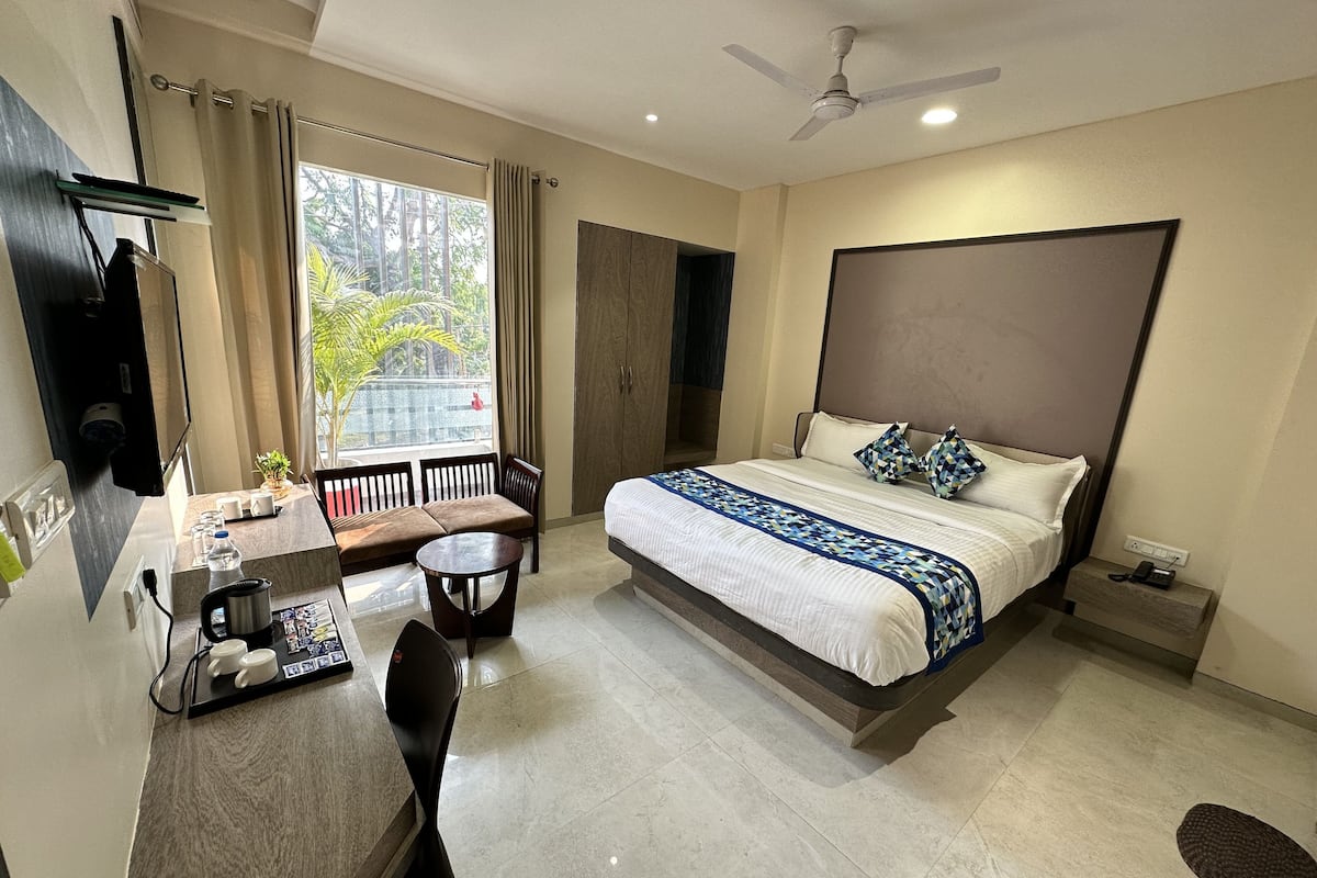A modern hotel room with a bed, desk, chair, and a view of greenery outside.
