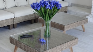 Una sala de estar con un sofá color beige, dos mesas auxiliares de mimbre y un jarrón con flores azules.