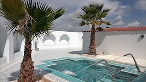 Una piscina con plataforma de baldosas, rodeada de palmeras y un edificio blanco con arcos.