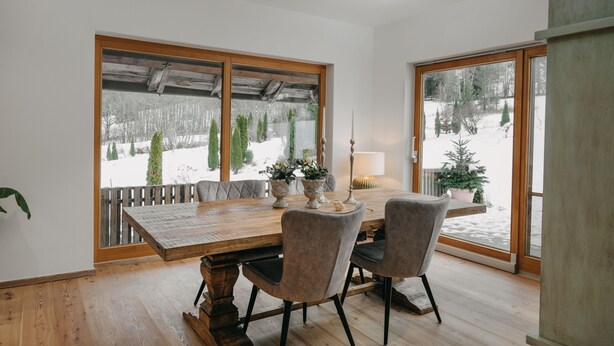 A dining area with a wooden table and chairs, large windows with a view of a snowy landscape, and a potted plant on the windowsill. - Apartment with 180sqm - in a former quiet farmhouse (Weitnau)