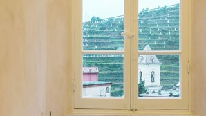 Ein Zimmer mit Fensterblick auf einen terrassierten Weinberg und ein kleines Gebäude. Ein Tisch mit Obstschale und ein Stuhl sind vorhanden.