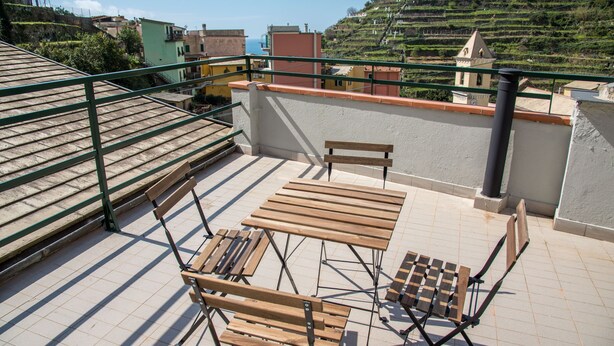 Eine Dachterrasse mit Holztisch und -stühlen, einem Metallgeländer und Blick auf terrassenförmig angelegte Felder und Gebäude.