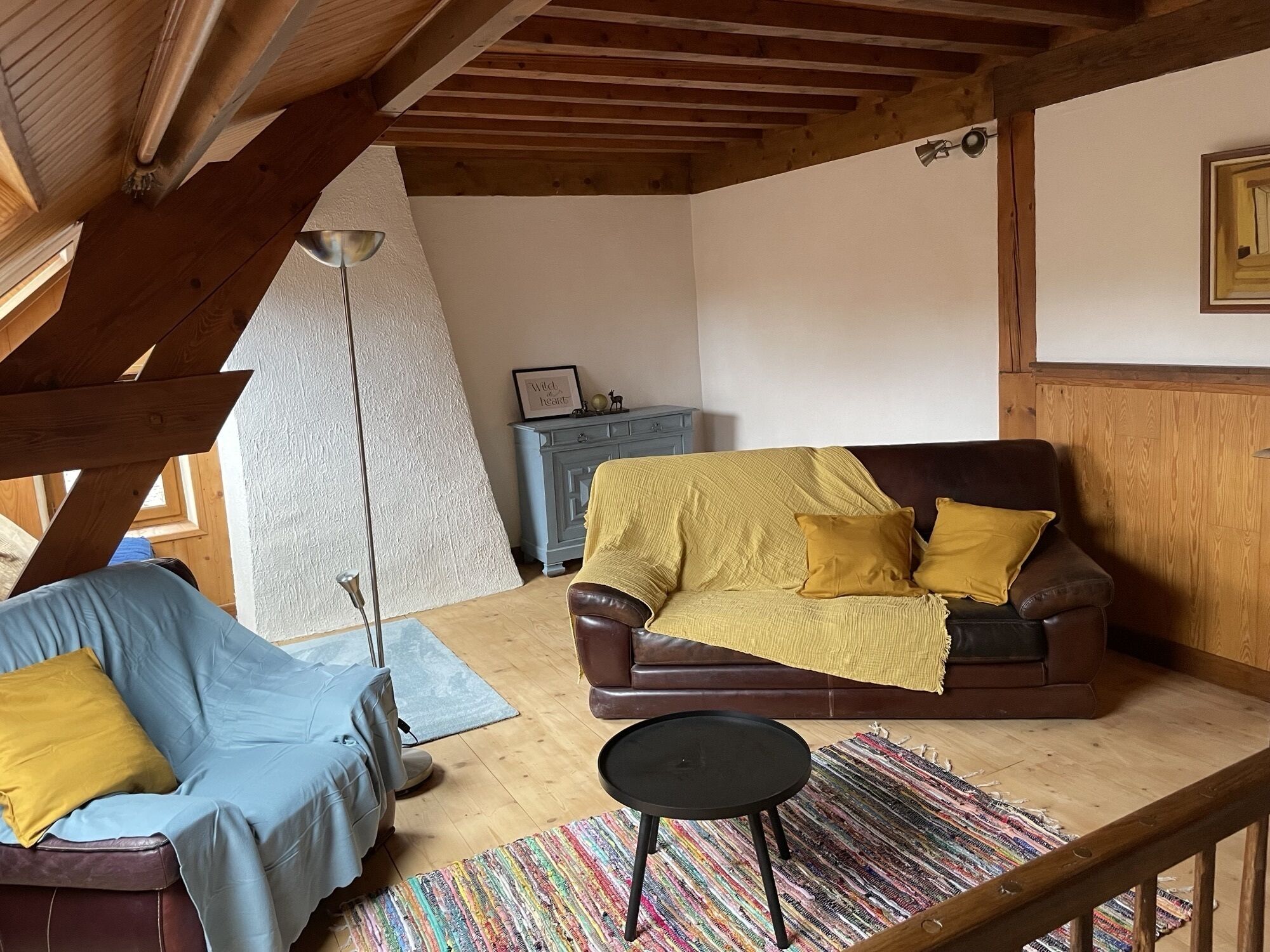 A cozy living room with a brown leather sofa, a blue armchair, and a small black table. A colorful striped rug is on the floor.