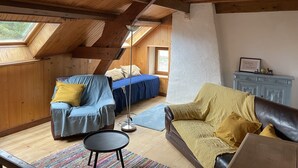 Une chambre mansardée chaleureuse avec des poutres en bois, un plafond en pente et un puits de lumière. On y trouve un lit avec une literie bleue et une fenêtre, une chaise bleue avec un coussin jaune, et un canapé en cuir marron avec un plaid jaune.