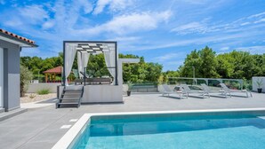 Un espace piscine extérieur moderne, avec un ciel bleu dégagé, un coin salon abrité et une piscine aux bords nets.