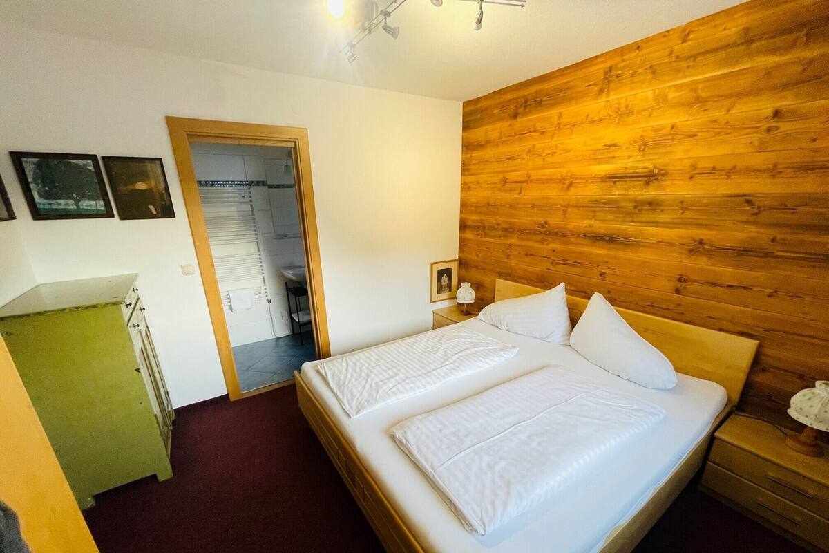 A hotel room with a wooden headboard, a bed with white linens, a green desk, and a bathroom visible through an open door.