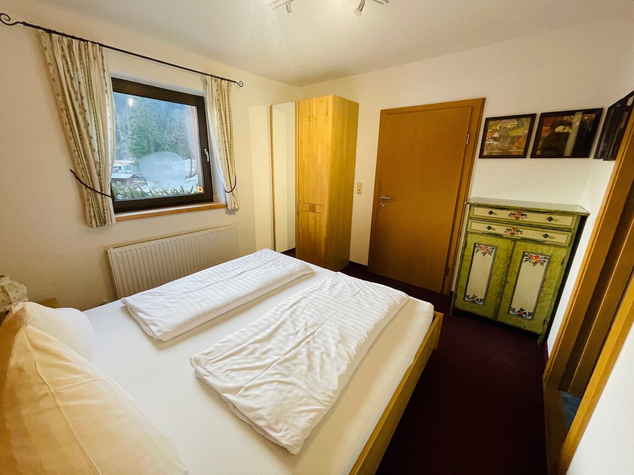 A bedroom with a bed, a window with curtains, a radiator, a wardrobe, and a dresser.