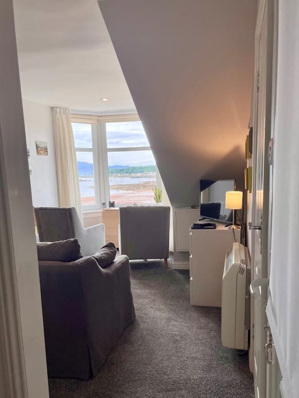 A room with a view of a beach, a desk with a lamp, a chair, and a bed.