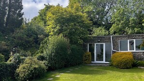 A garden with a wooden table and benches, surrounded by lush greenery and a stone building with glass doors. - Millers cottage, a picturesque semi detached cottage with private garden (Woolfardisworthy)