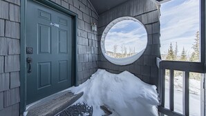 Exterior de fusta amb una porta verda, un porxo cobert de neu i una finestra circular que mostra vistes als arbres.