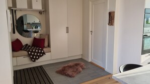 Ein kompakter Wohnraum mit Küchenzeile, einer Sitzecke mit Sofa und Kissen, einem flauschigen Haustier auf dem Boden und einer Tür zu einem weiteren Raum.