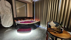 A modern bathroom with a marble vanity, a freestanding bathtub with rose petals, candles, and a wooden side table with a bottle and glass. - Apricot Motera  (Ahmedabad)
