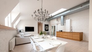 A modern dining area with a white table set for six, surrounded by white chairs. A chandelier hangs from the ceiling, and a wooden cabinet is visible in the background. - 2ndhomes Luminous 2BR Loft House w Sauna (Helsinki)