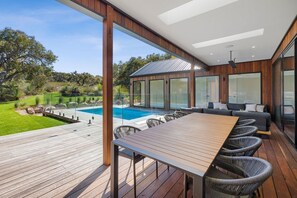 Una terraza moderna al aire libre con mesa y sillas de madera, una piscina y un área de estar cubierta con paredes de vidrio.