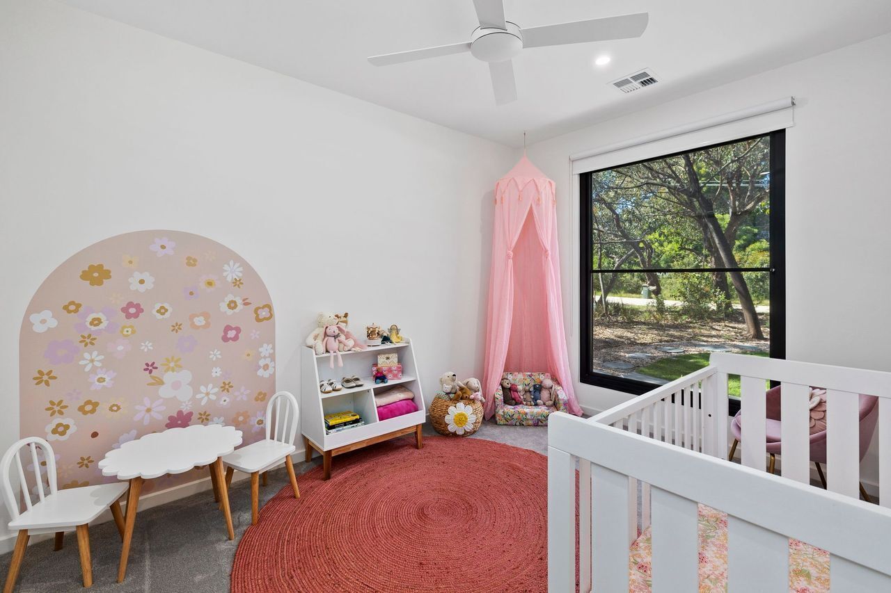 Une chambre d’enfant avec un berceau, une chaise blanche, une petite table, une bibliothèque et une grande fenêtre donnant sur des arbres.