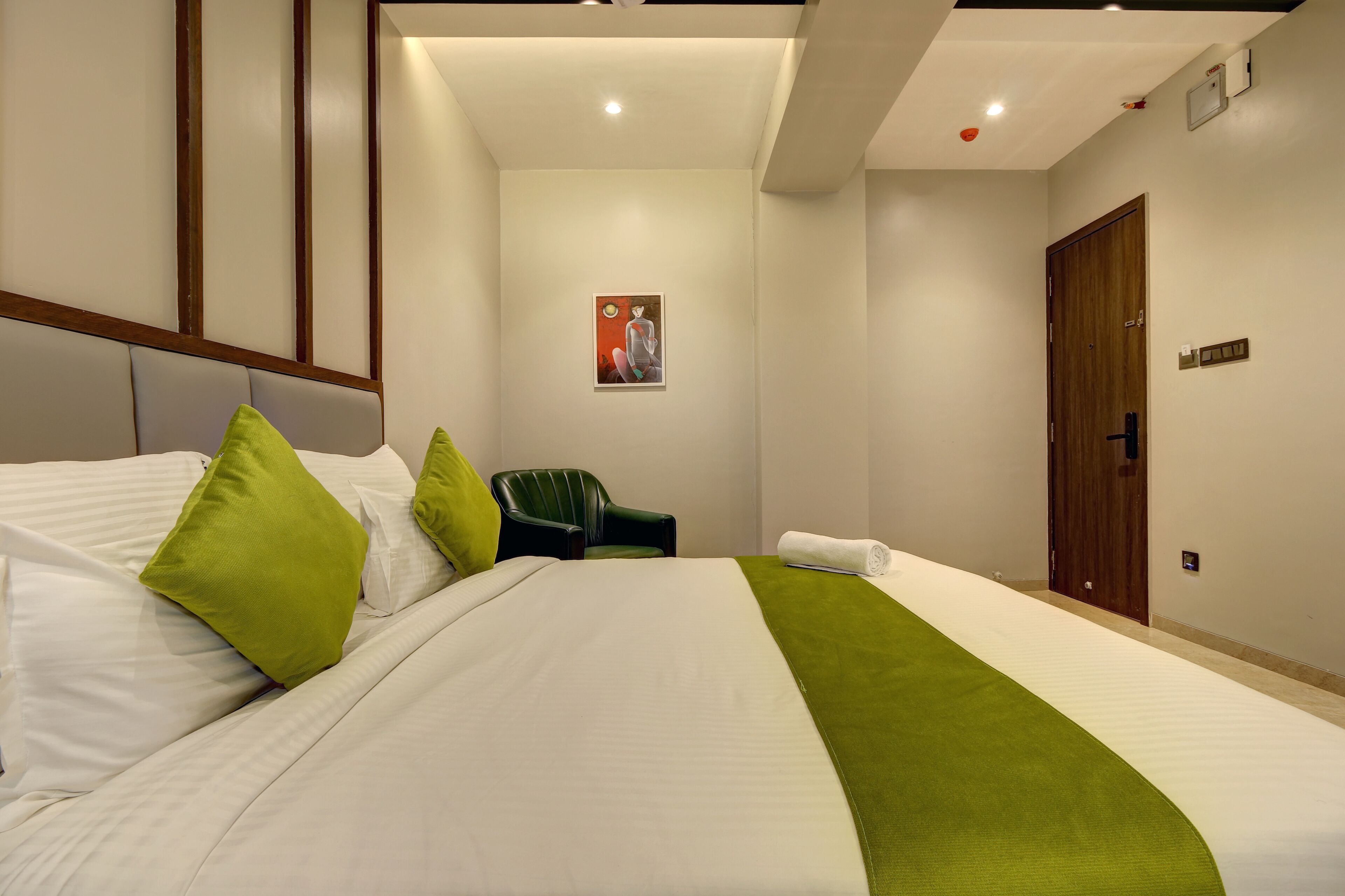 A hotel room with a large bed, green pillows, a green throw, a chair, and a door.