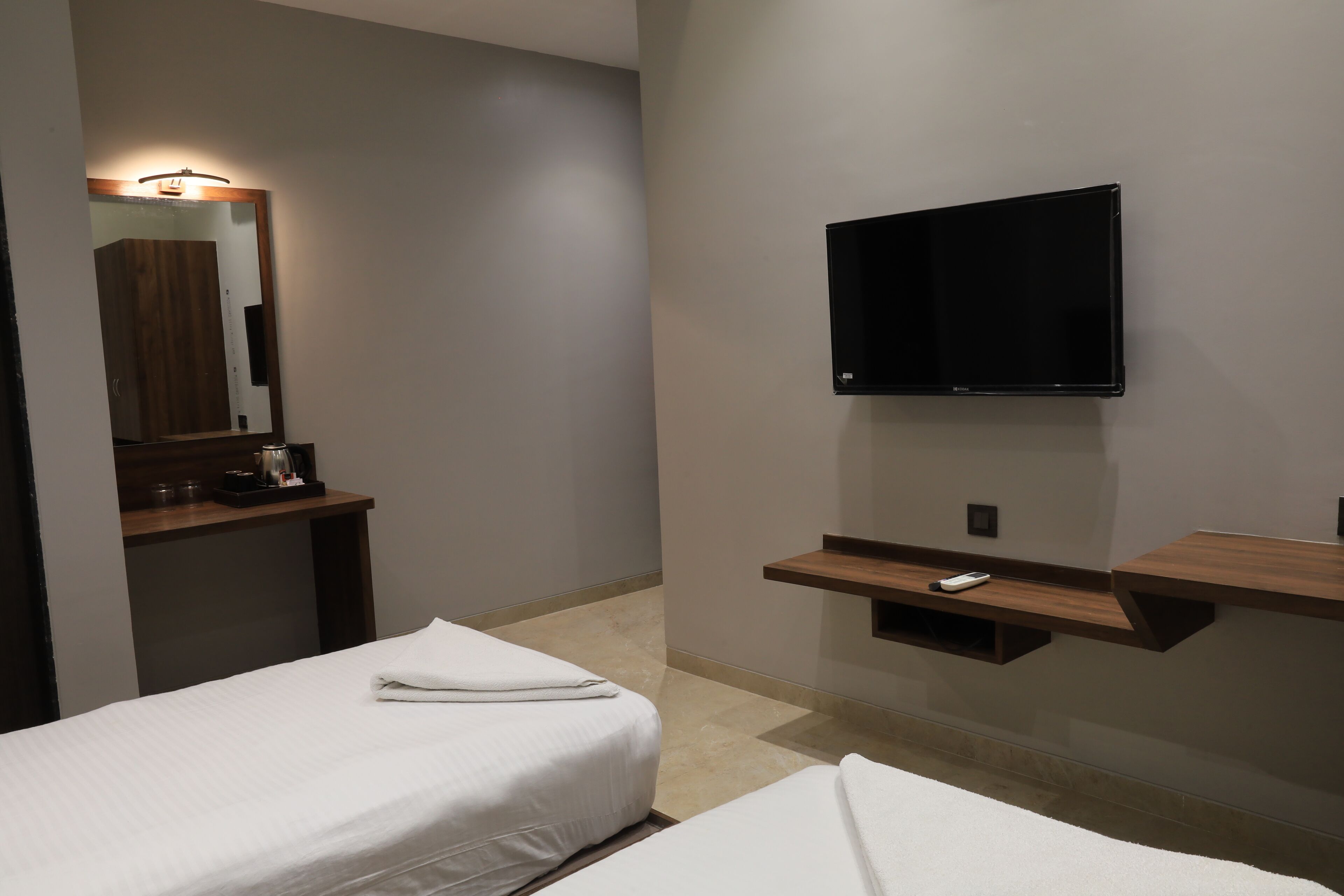 Habitación de hotel con dos camas, un televisor montado en la pared, un espejo sobre una repisa de madera y una pequeña mesa con un control remoto.
