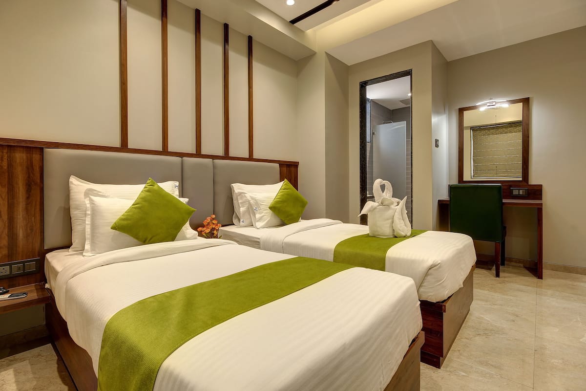 A hotel room with two beds, each with white bedding and green accents, a wooden headboard, a desk with a chair, and a mirror on the wall.