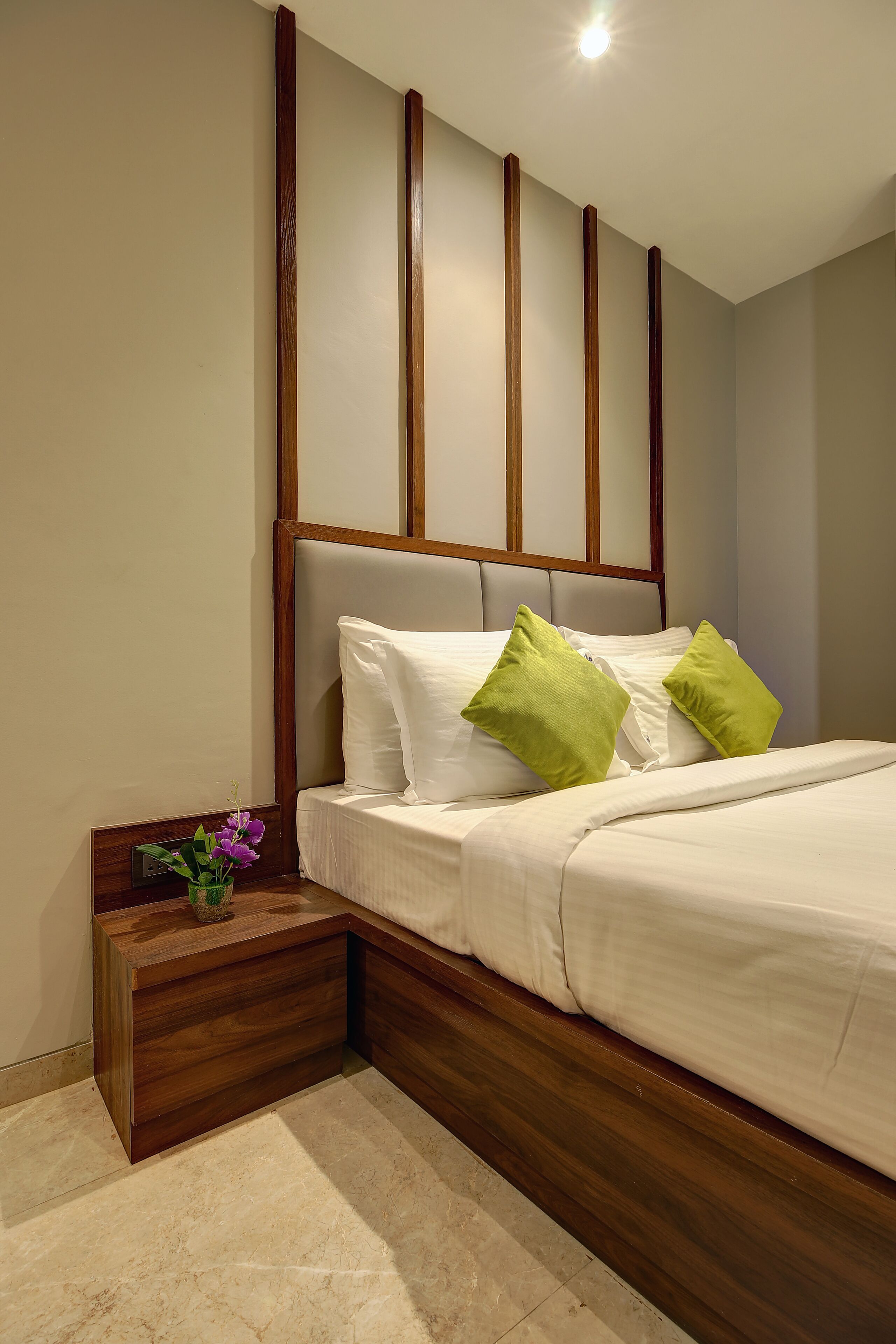 A neatly made bed with white linens and two green pillows, a wooden nightstand with a vase of purple flowers, and a headboard with vertical wooden panels.