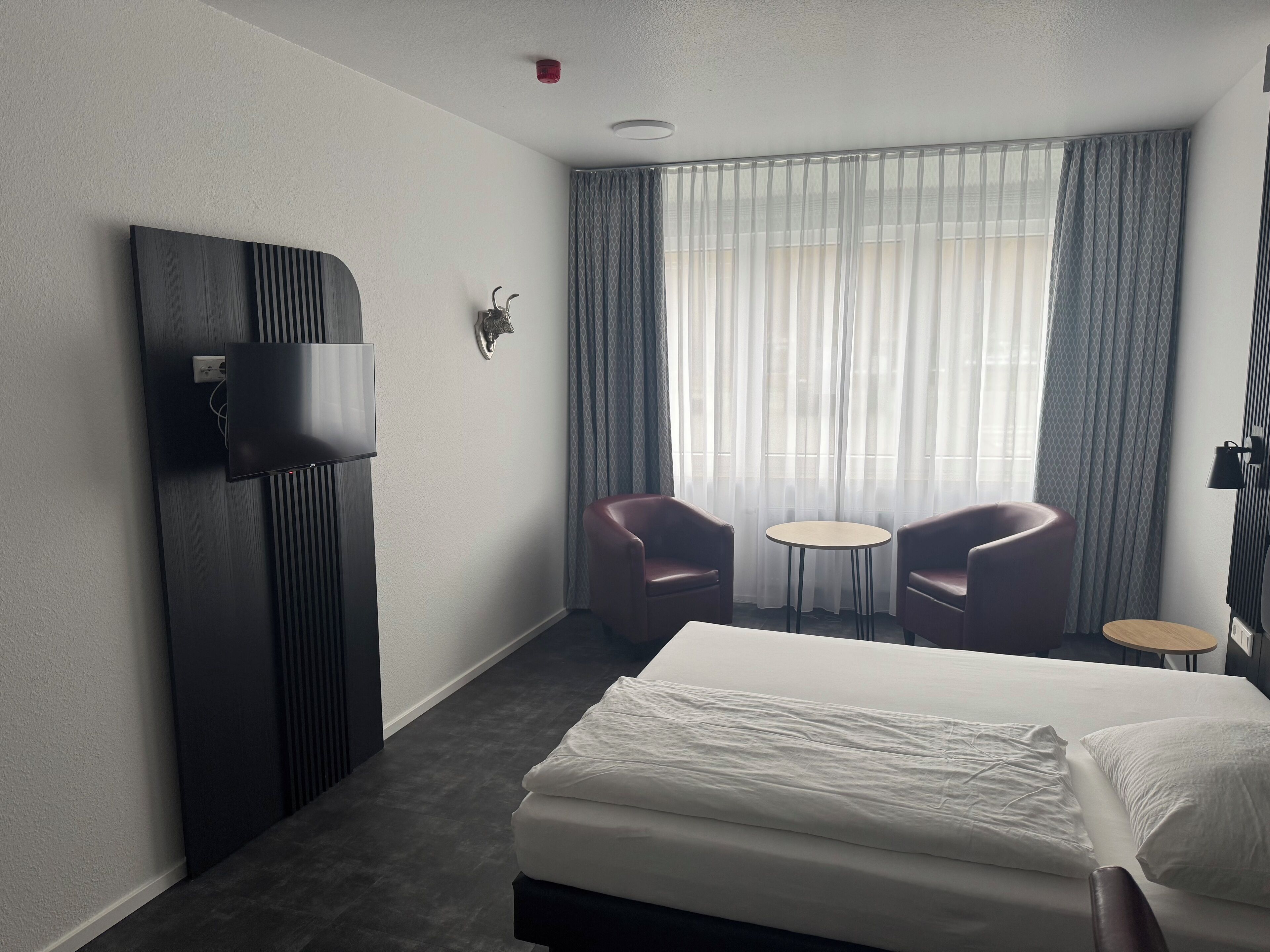 A hotel room with a bed, two armchairs, a small table, a TV mounted on a dark panel, and a window with sheer curtains.