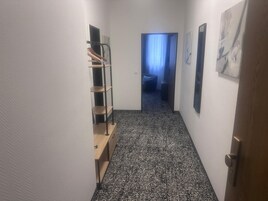 Um corredor estreito com carpete estampado em preto e branco, uma escrivaninha de madeira com gavetas, uma estante de metal e uma porta que dá para outro quarto.