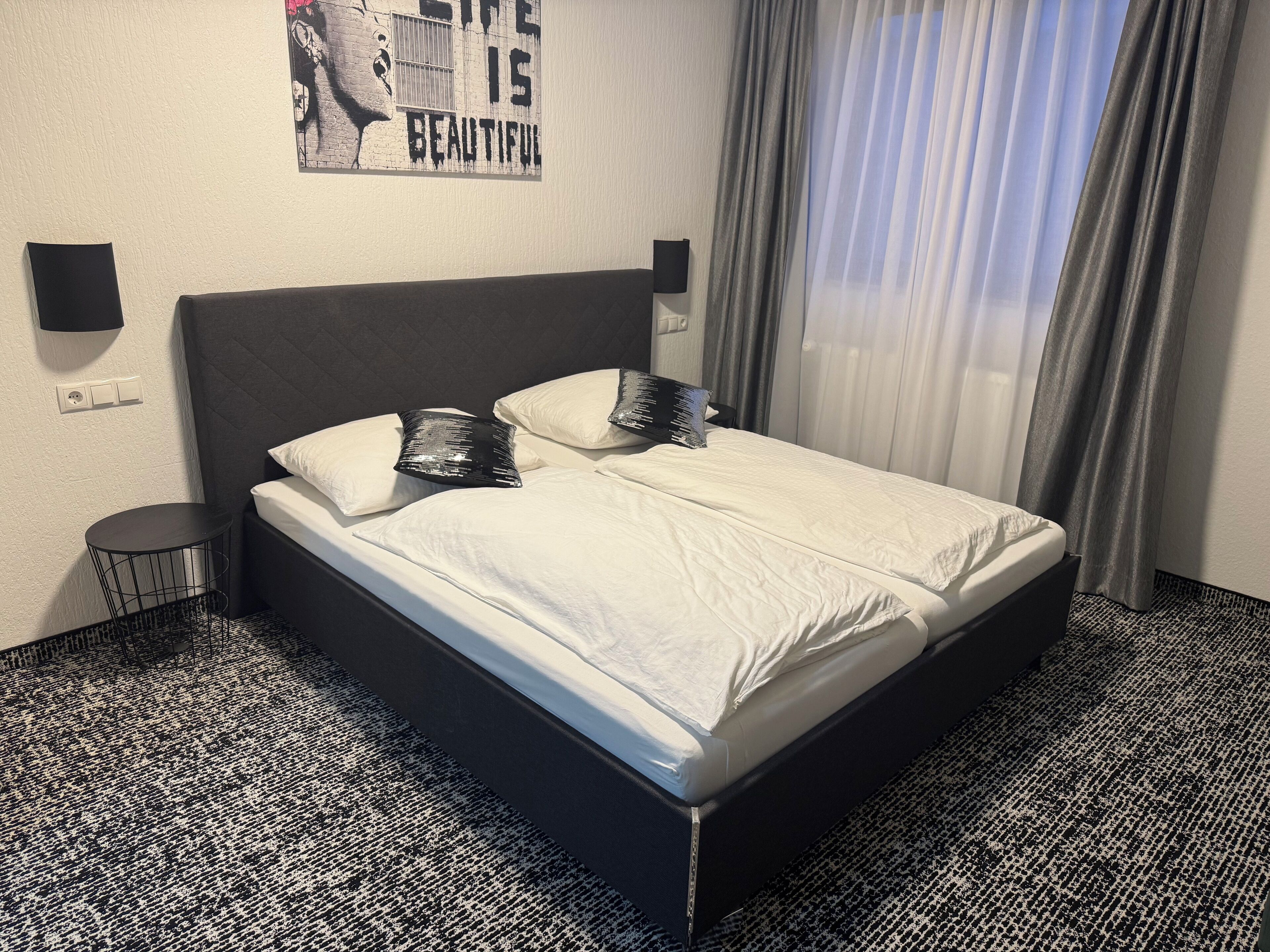 Habitación de hotel con cabecera negra, dos camas blancas, alfombra con estampado blanco y negro, una mesita redonda y una pared con un cartel.