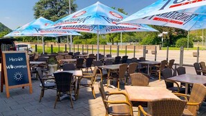 An outdoor seating area with wicker chairs and tables, covered by branded umbrellas, and a sign that reads "Herzlich Willkommen". - Panorama Hotel Rheinkrone (Andernach)