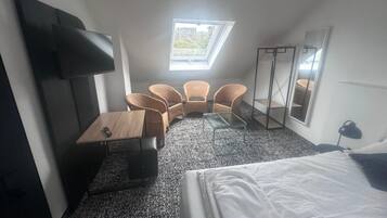 A hotel room with a bed, a TV mounted on the wall, a small table, two wicker chairs, a standing mirror, and a window with a view of greenery.