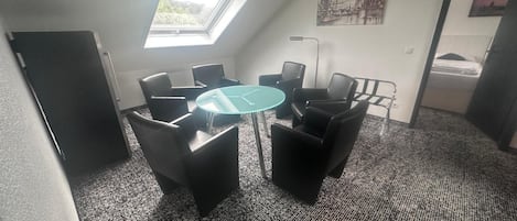 Un salón de conferencias con una mesa redonda de cristal, seis sillas negras, un tragaluz y un cuadro de paisaje urbano en la pared.