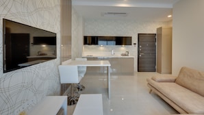 A modern living room with a flat-screen TV, a white sofa, a dining table, and a kitchen area in the background.