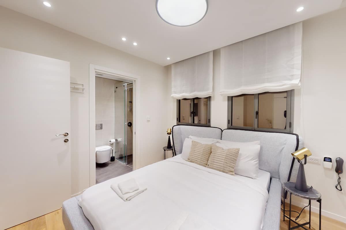 A bedroom with a bed, bedside lamps, a bedside table, a window with curtains, and a door leading to a bathroom.