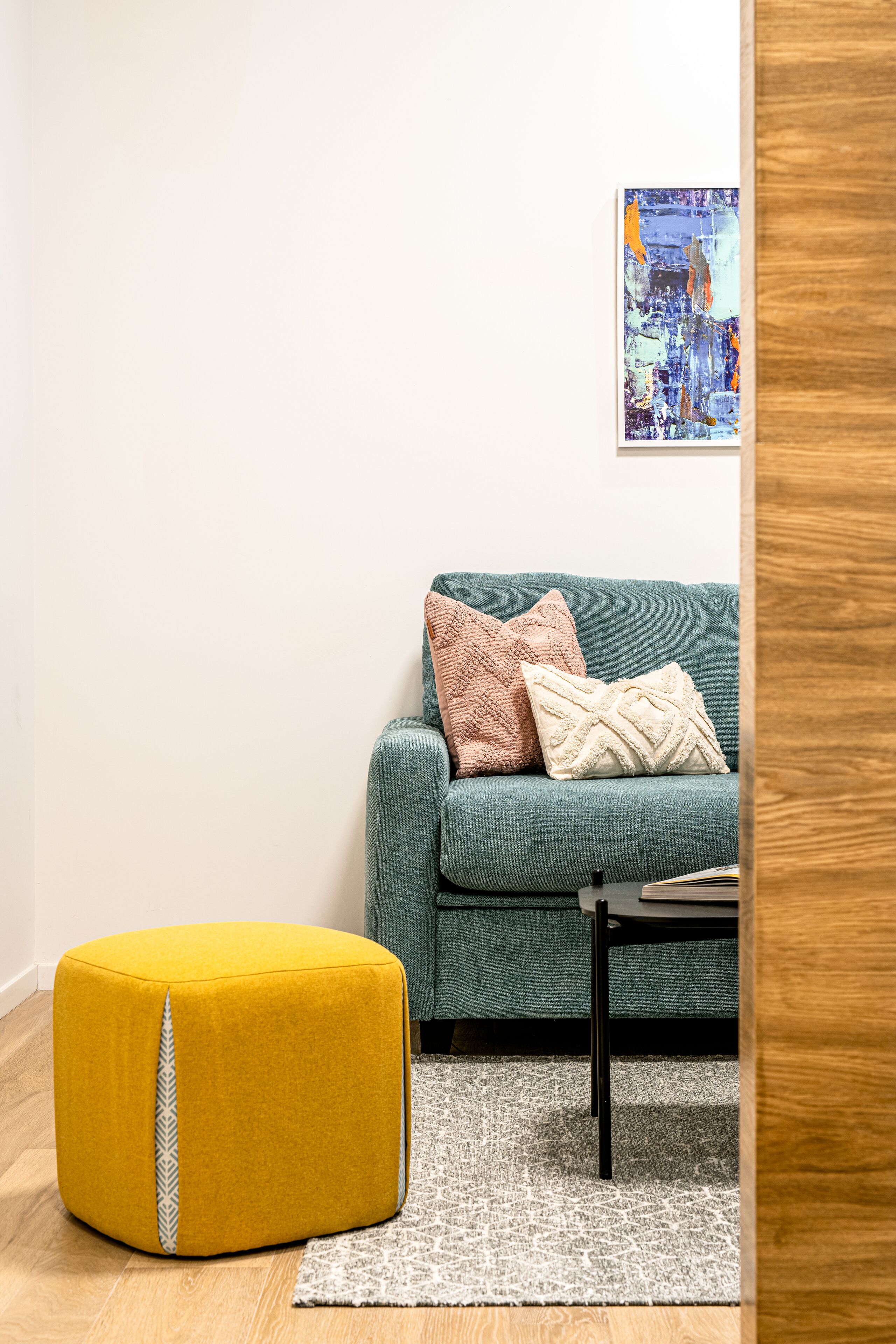 A living room with a teal sofa, a yellow ottoman, and a black coffee table.
