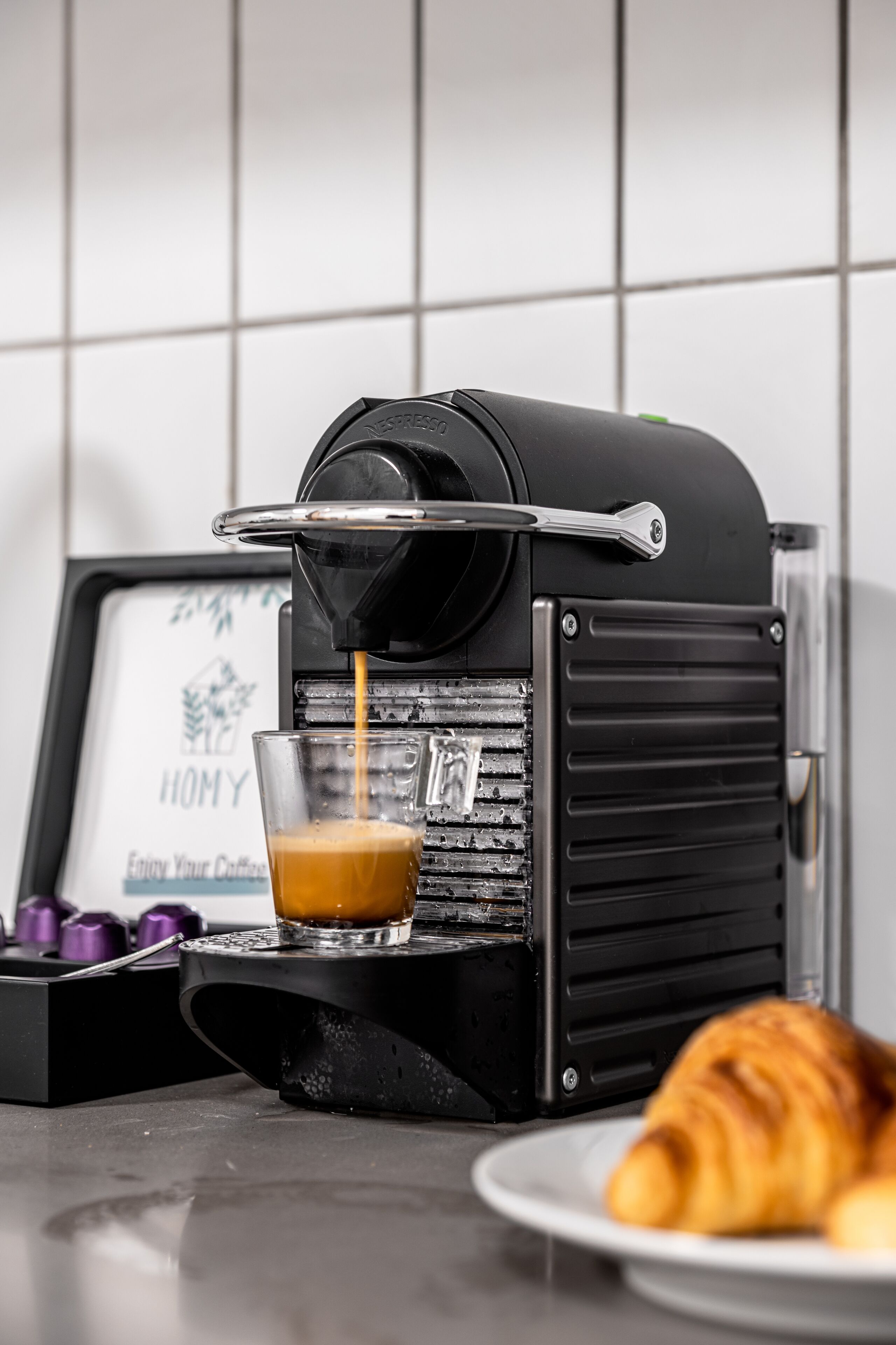 A coffee machine dispensing espresso into a glass, with a croissant on a plate beside it.