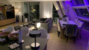 A modern living room with a sofa, dining table, and chairs. There is a TV, a lamp, and a plant. - Stunning city penthouse with 360-degree skyline views (London)