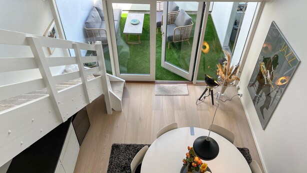 Un intérieur moderne avec un escalier, un coin repas doté d’une table ronde et une vue sur un jardin extérieur, visible à travers des portes vitrées.