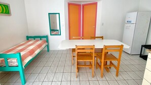Una habitación pequeña y luminosa con una cama individual, una mesa con sillas, un refrigerador y una puerta con paneles rosa y naranja.