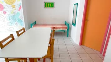 A room with a long white table and wooden chairs, a ceiling fan, a green exit sign, and a door with a pink frame.