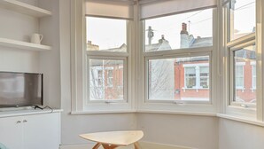 Habitación de esquina con ventanas amplias, un banco de madera, un televisor sobre un mueble blanco y vista a un edificio de ladrillo rojo.