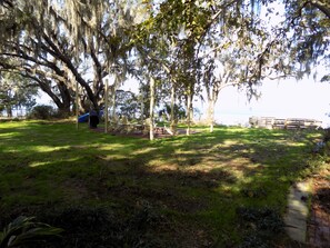 A grassy outdoor area with a wooden deck and chairs, a grill, and a view of a body of water. - Secluded Waterfront Home on the Bay (Pensacola)